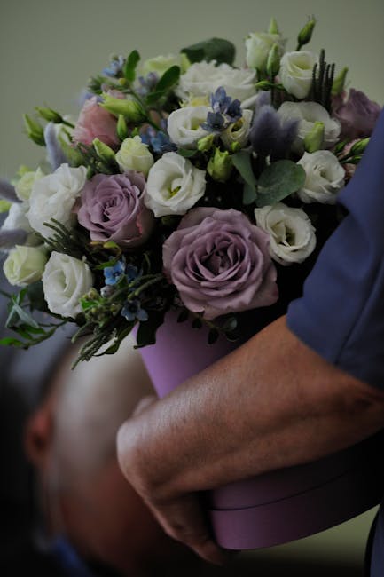 A person holding a bouquets of fresh flowers, featuring lavender-hued roses, white lisianthus, and small blue blossoms, accented with green foliage. The arrangement is bound with a soft purple wrappin