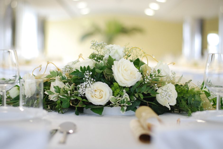 A floral arrangement featuring white roses, calla lilies, and baby's breath, accented with green foliage such as fern leaves, arranged in a low, elongated display on a white tablecloth. The bouquet is