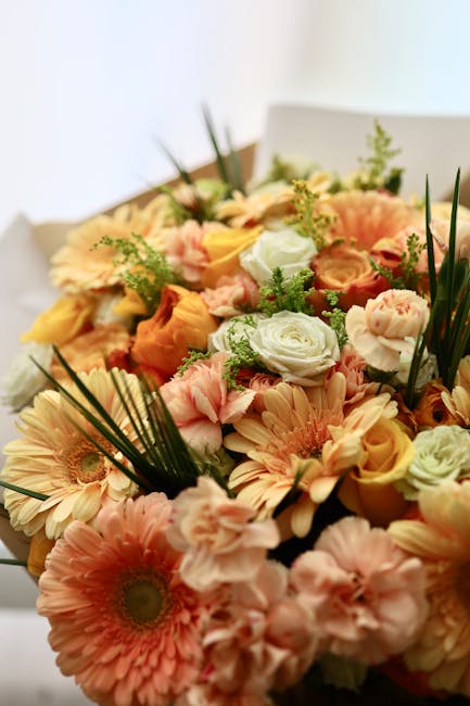 A vibrant floral bouquet featuring a mix of peach, cream, and light pink roses, along with peach and white gerbera daisies and delicate pink lisianthus, all accented by lush greenery and long, slender