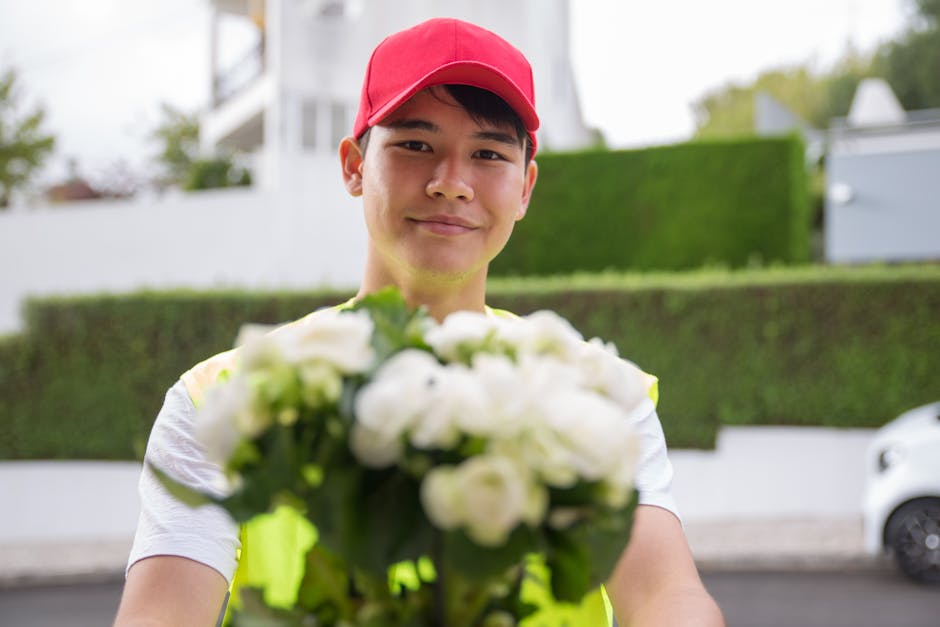 A delivery person wearing a red cap and a reflective yellow vest is holding out a bouquet of white flowers towards the camera, standing outdoors near a residential area with a background of green bush