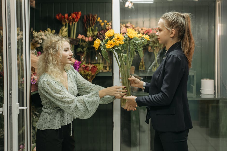 A florist with curly blonde hair and a patterned blouse is handing a bouquet of bright yellow sunflowers, arranged with green foliage, to a customer in a floral shop. The customer, with long hair tied