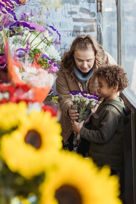 A woman and a young boy are inside a floral shop near a large window, engaging in a flower delivery or floral gift occasion. The woman, with light brown wavy hair and a beige coat, is handing a small 