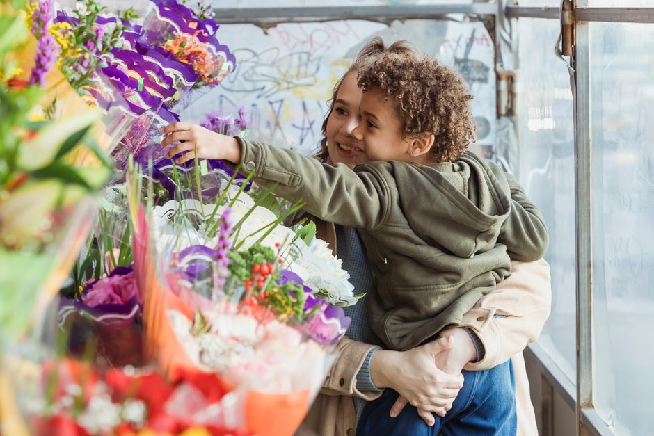 A young child with curly hair and a joyful expression is being held by an adult in a floral shop, reaching out to select a vibrant bouquet of fresh flowers. The bouquet features an array of colorful b
